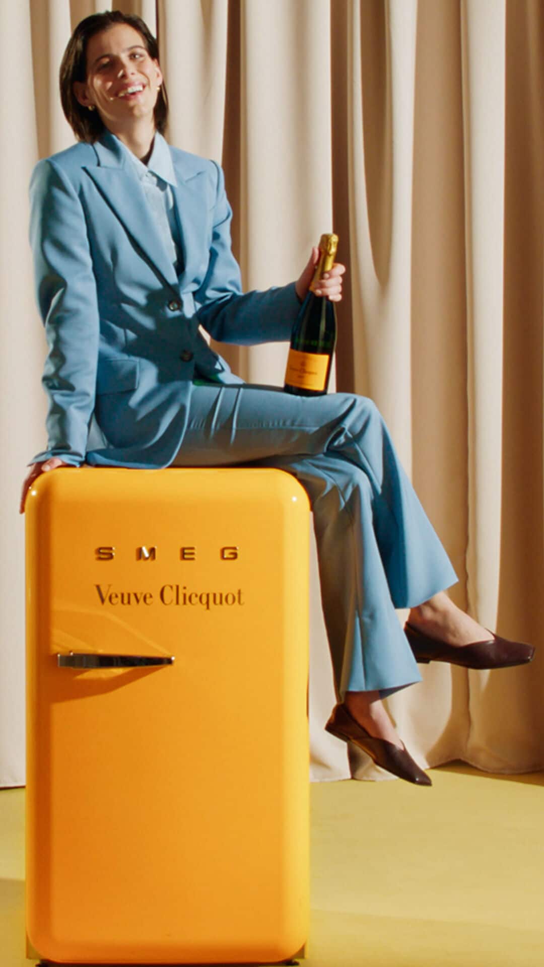 Veuve Clicquot Brut Yellow Label showcased by a Lady sitting over a Veuve Clicquot yellow colored mini fridge