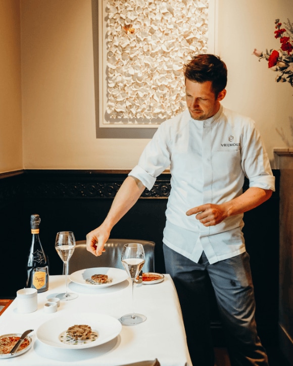 Michaël Vrijmoed in his restaurant serving two plates, with glasses of champagne and a bottle of La Grande Dame 2015