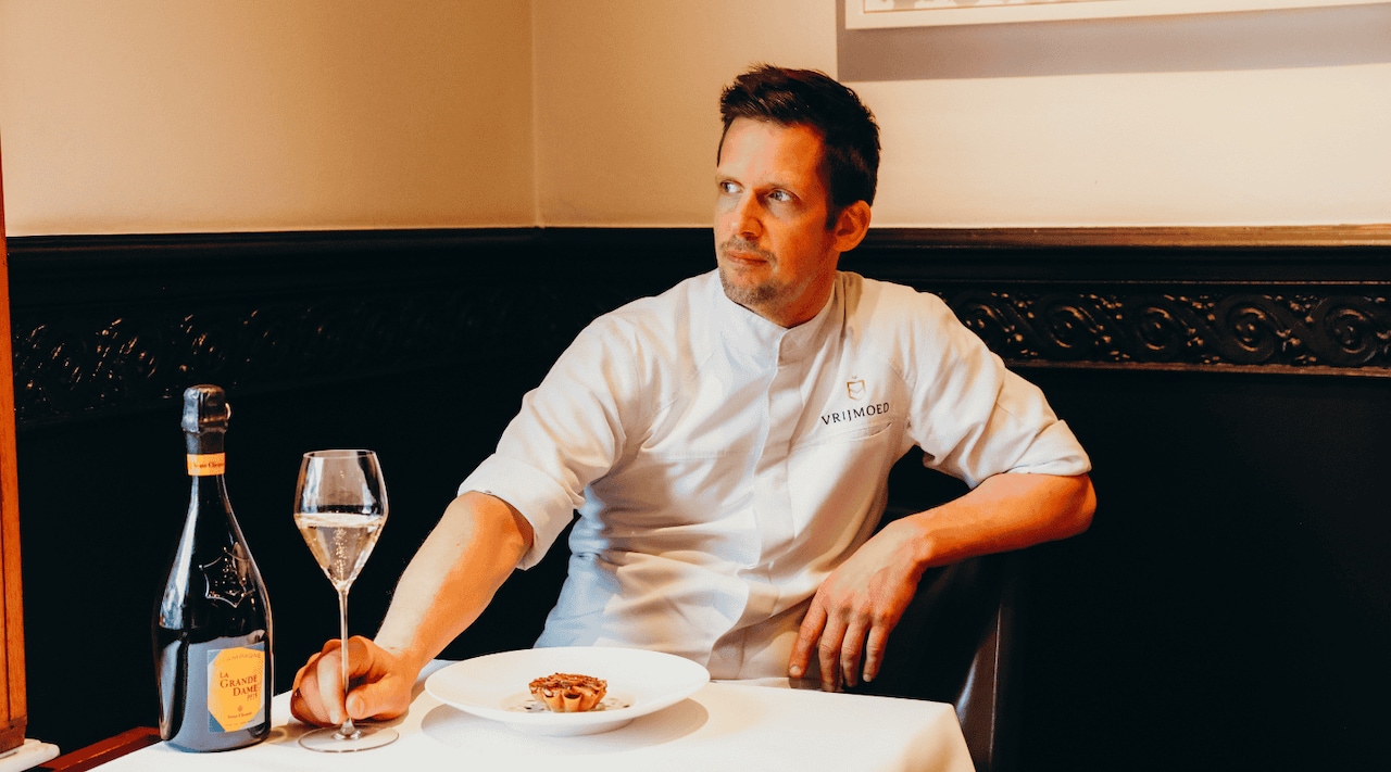 Michaël Vrijmoed in his restaurant with a dish, a glass of champagne and a bottle of La Grande Dame 2015