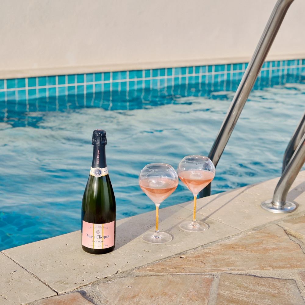 Bottle of RICH Rosé champagne with two glasses by a pool Bottle of RICH Rosé champagne with two glasses by a pool
